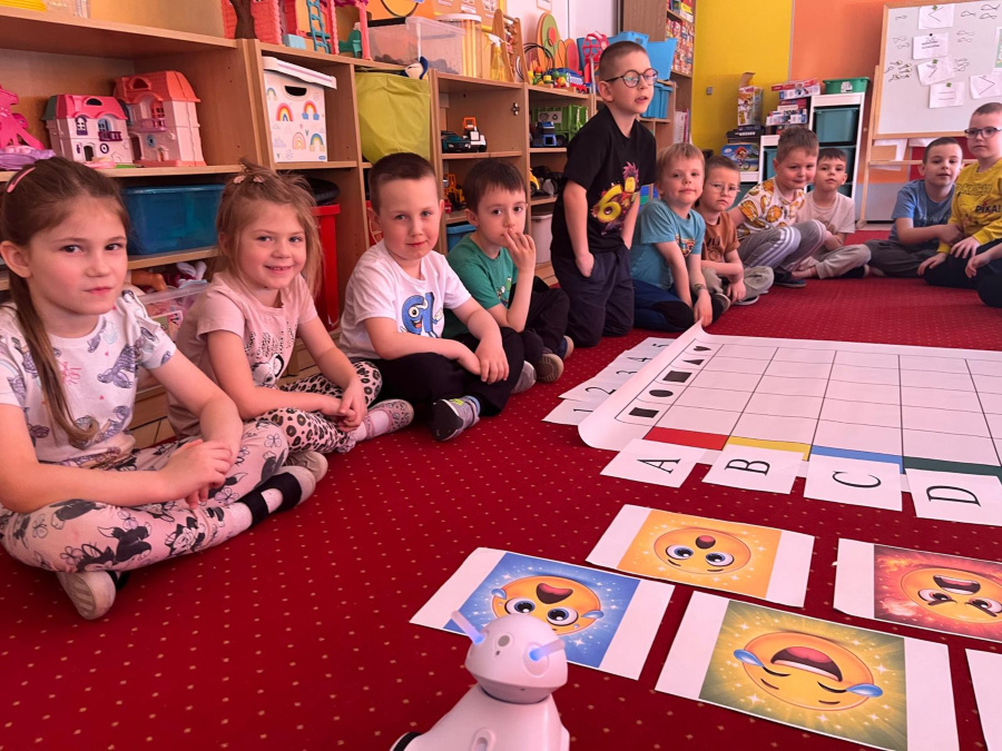 Grupa dzieci siedzi w półkolu na czerwonym dywanie. Przed nimi leży duża mata podzielona na kratki oraz kartki z literami A, B, C i D. Na podłodze ułożone są także karty z kolorowymi emotikonami 