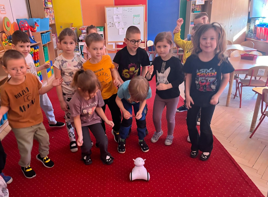 Grupa dzieci stoi na czerwonym dywanie i obserwuje małego białego robota znajdującego się na podłodze. Niektóre dzieci podskakują i unoszą ręce reagując na jego ruch