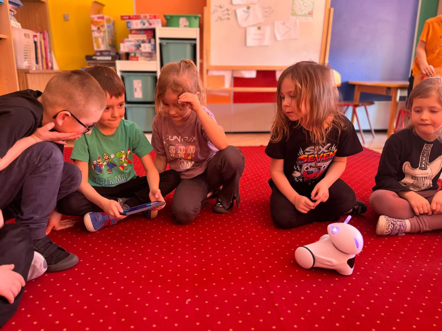 Kilka dzieci klęczy na czerwonym dywanie i obserwuje małego białego robota z podświetlonymi elementami. Jedno z dzieci trzyma w rękach telefon którym steruje robotem. W tle widoczne są półki z zabawkam