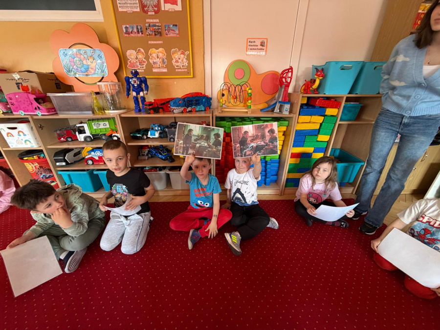Dzieci siedzą na czerwonym dywanie w sali przedszkolnej. Dwóch chłopców trzyma ilustracje a pozostali uczestnicy obserwują zajęcia. 