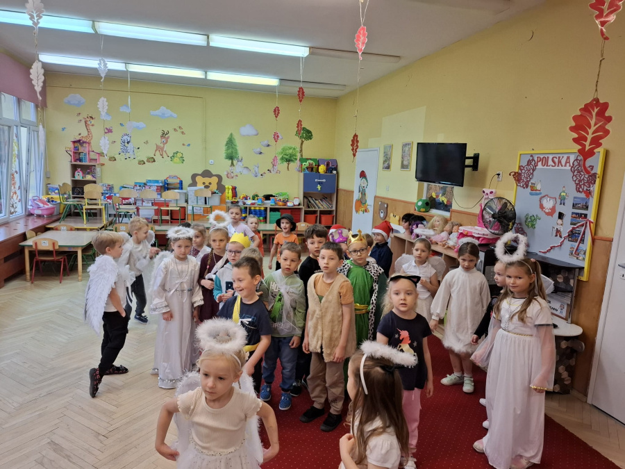 Dzieci stoją w sali ubrane w kostiumy przedstawiające anioły, świętych i postacie biblijne. Wnętrze ozdobione jest jesiennymi liśćmi zawieszonymi pod sufitem