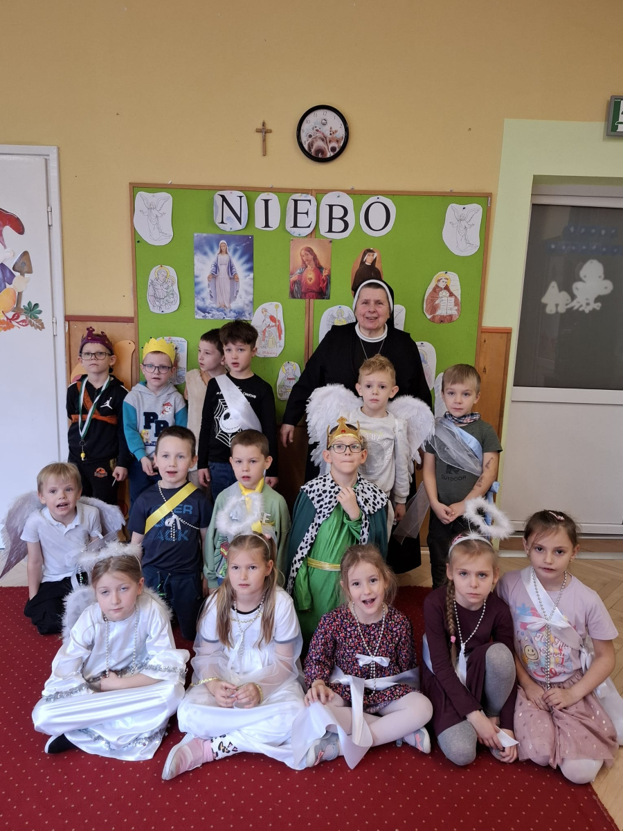 Grupa dzieci pozuje do wspólnego zdjęcia z siostrą zakonną. Dzieci mają na sobie przebrania przedstawiające anioły świętych i postacie królewskie