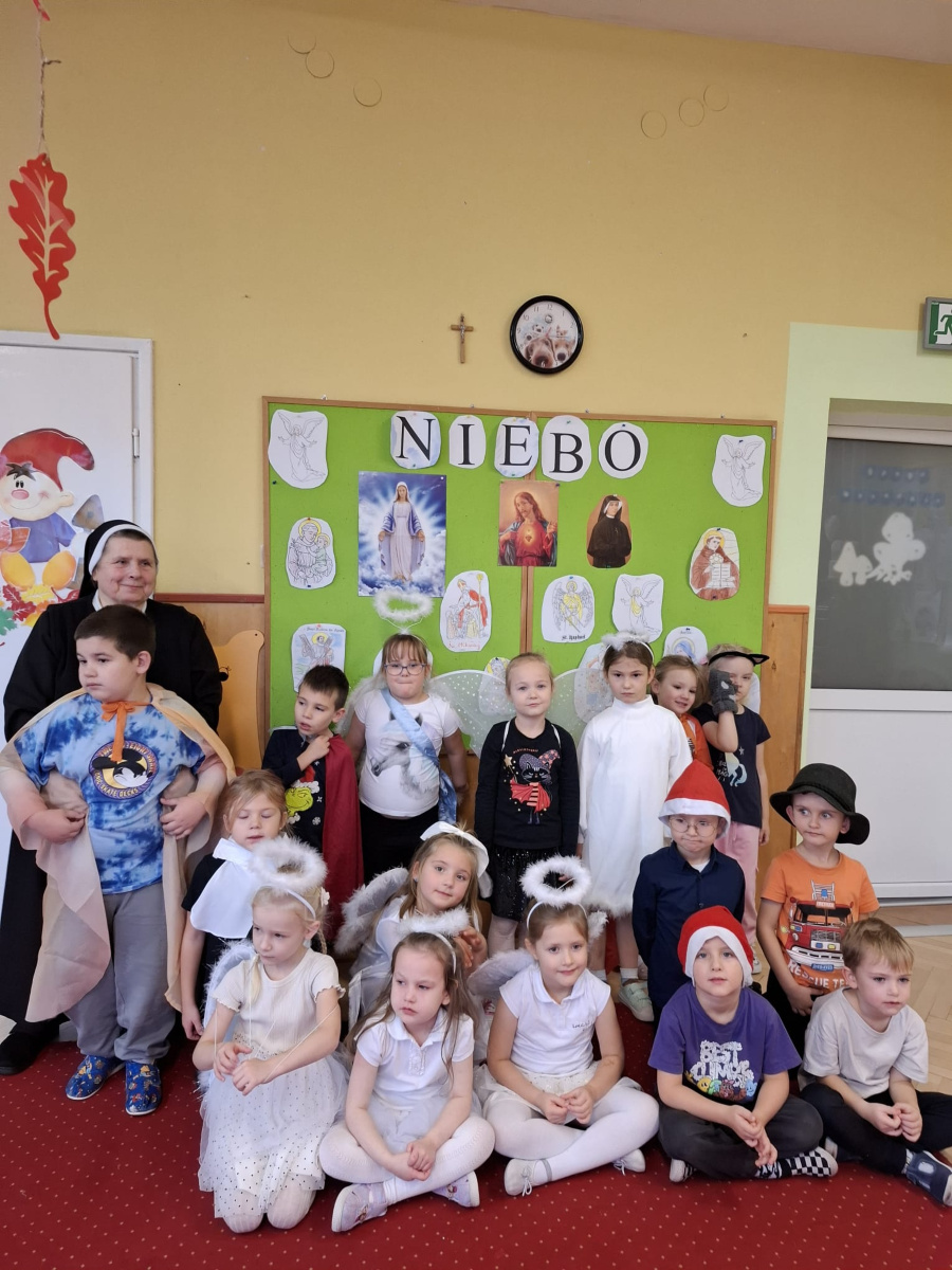 Grupa dzieci pozuje do wspólnego zdjęcia z siostrą zakonną. Dzieci ubrane są w przebrania przedstawiające anioły i świętych. Tło stanowi zielona tablica z napisem miebo