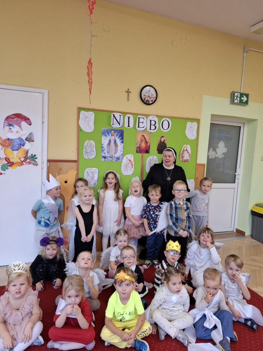 Grupa dzieci pozuje do wspólnego zdjęcia z siostrą zakonną. Dzieci ubrane są w stroje aniołki królów i świętych. W tle znajduje tablica z ilustracjami postaci religijnych i aniołó