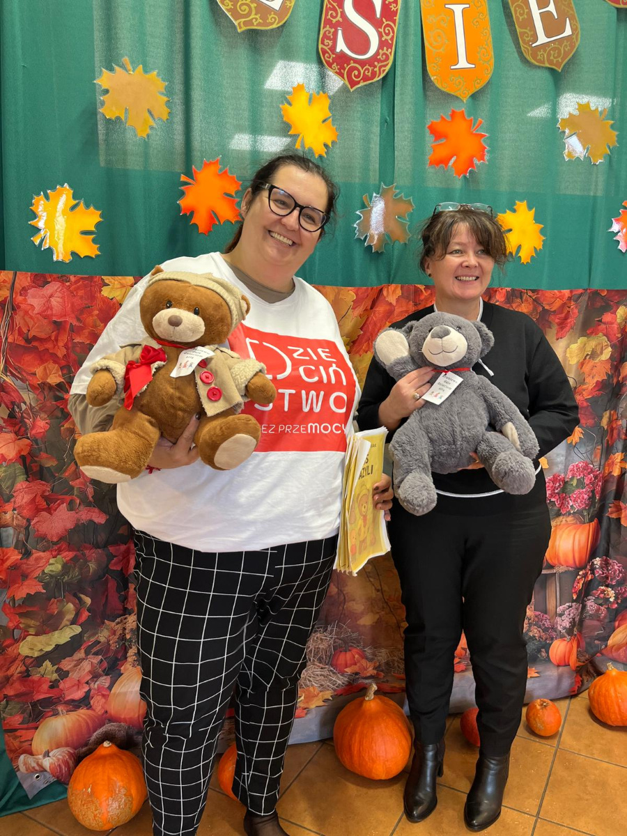 Dwie kobiety stoją na tle jesiennej dekoracji z kolorowych liści i napisu JESIEŃ. Każda z nich trzyma pluszowego misia. U ich stóp leżą dynie
