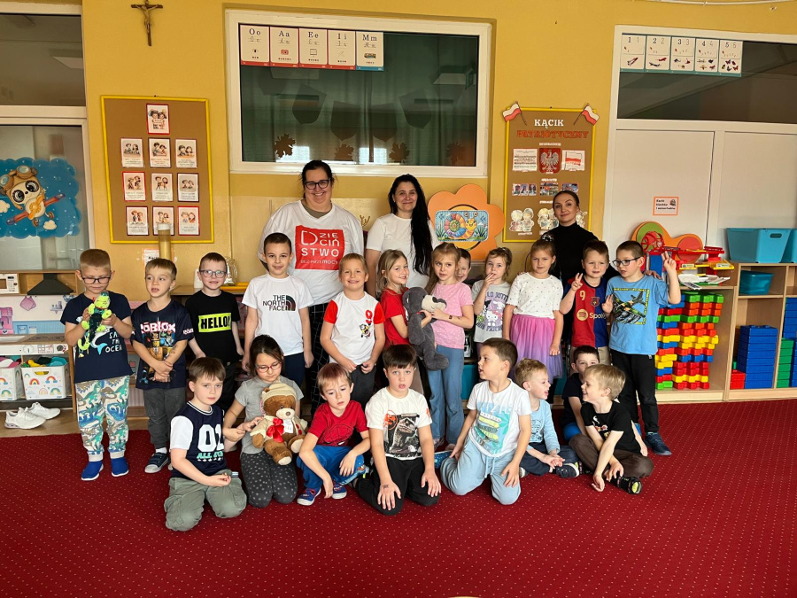 Grupa dzieci pozuje wspólnie z trzema nauczycielkami w sali przedszkolnej. Niektóre dzieci trzymają pluszowe misie. W tle widać kolorowe półki z zabawkami, plansze edukacyjne oraz dekoracje