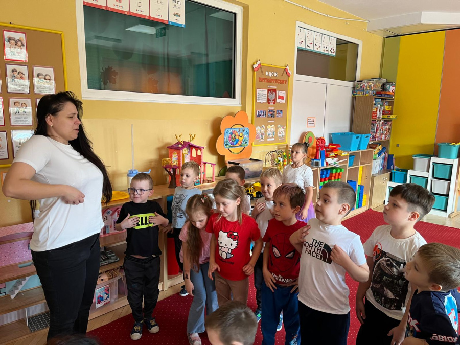 Kobieta stoi naprzeciw grupy dzieci i pokazuje gest a dzieci naśladują jej ruchy. W pomieszczeniu widać kolorowe meble zabawki oraz plansze dydaktyczne na ścianach