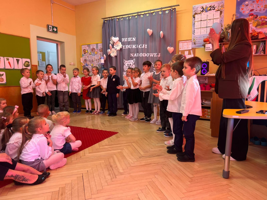 Dzieci w odświętnych strojach stoją w półkolu. Przed nimi siedzą inne dzieci obserwujące przedstawienie a obok nauczycielka bije brawo