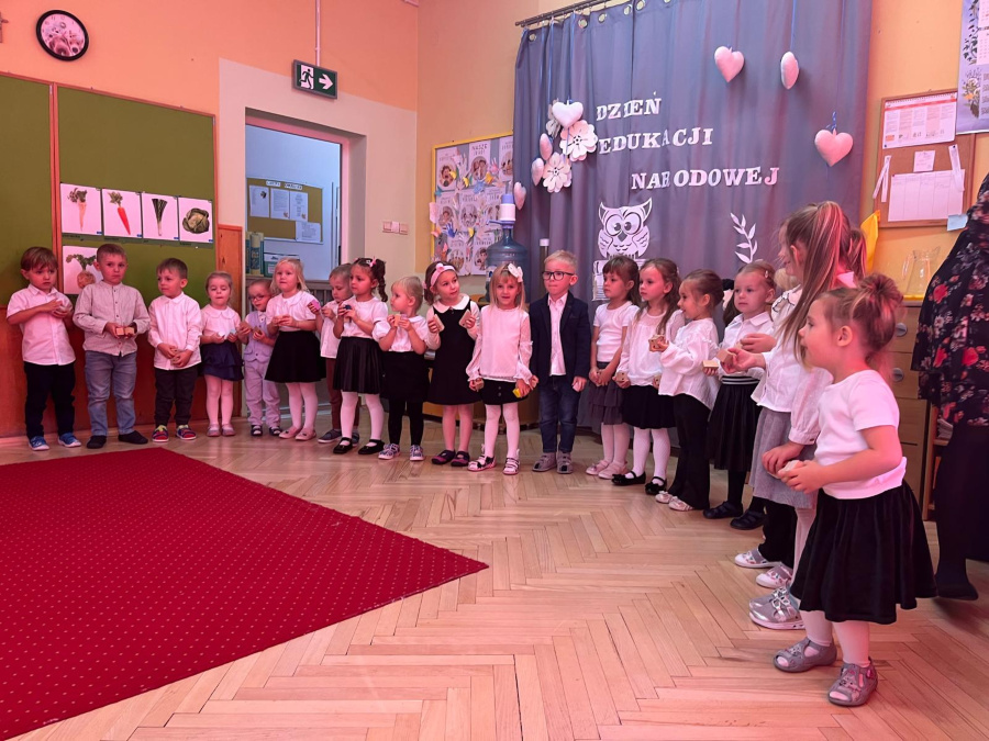 Grupa dzieci stoi w szeregu podczas występu z okazji Dnia Edukacji Narodowej. Dzieci ubrane są w białe bluzki koszule i ciemne spódniczki lub spodnie. Trzymają w dłoniach instrument