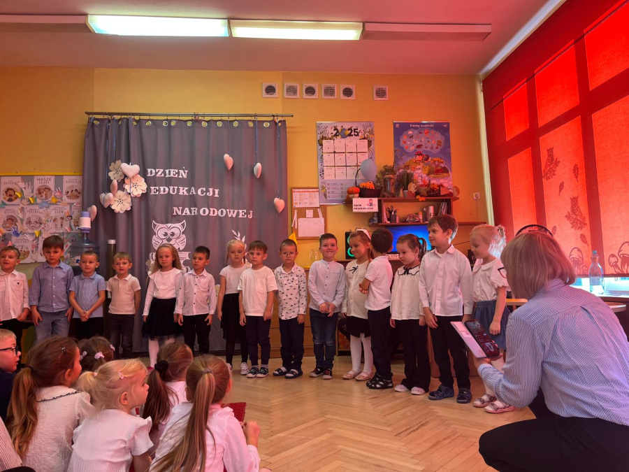 Grupa dzieci stoi w szeregu przed ozdobioną kotarą z napisem „Dzień Edukacji Narodowej”. Dzieci ubrane są w białe koszule i ciemne spodnie lub spódniczki. Przed nimi siedzą dzieci