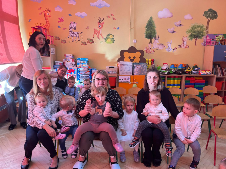 Nauczycielki i dzieci siedzą razem w sali ozdobionej kolorowymi ilustracjami zwierząt. Dzieci siedzą na kolanach dorosłych lub na małych krzesełkach niektóre uśmiechnięte inne poważ