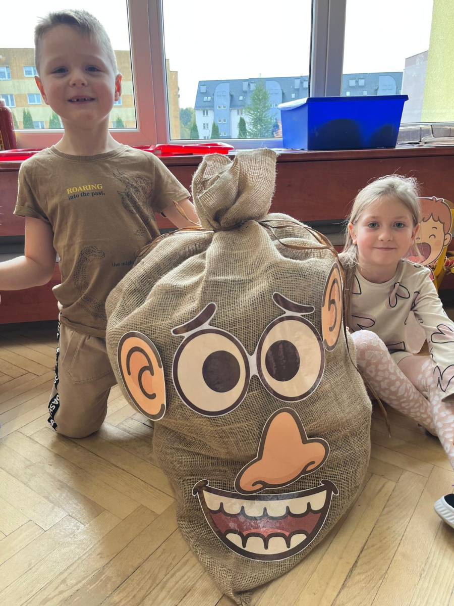Dwoje dzieci siedzi na podłodze obok dużego worka ozdobionego papierowymi elementami części twarzy. Chłopiec po lewej stronie ma krótkie włosy i beżową koszulkę