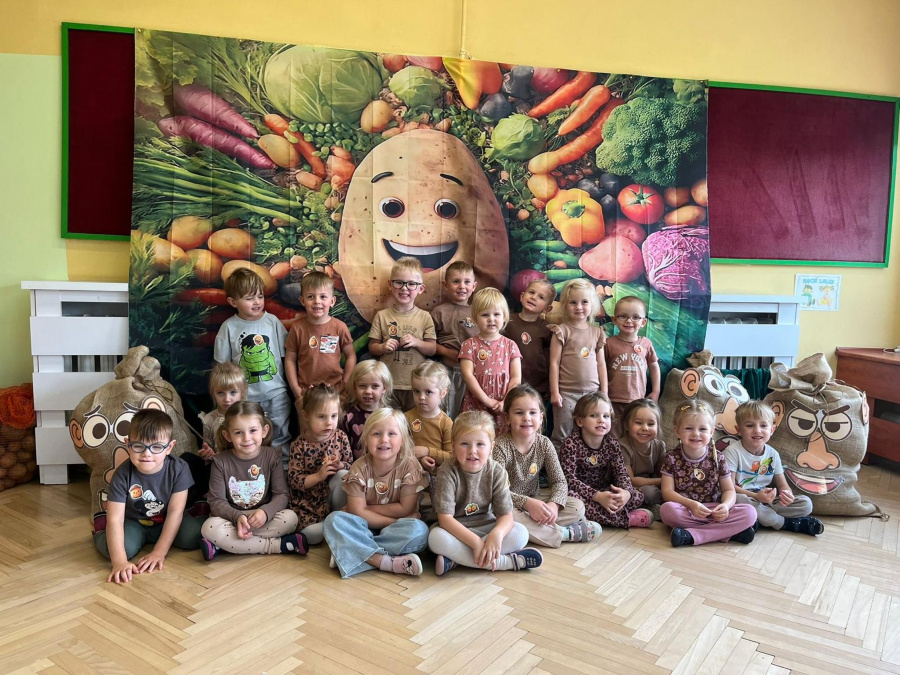 Grupa dzieci pozuje do wspólnego zdjęcia na tle dekoracji przedstawiającej uśmiechniętego ziemniaka otoczonego warzywami. Dzieci ubrane są w stroje w odcieniach brązu i beżu