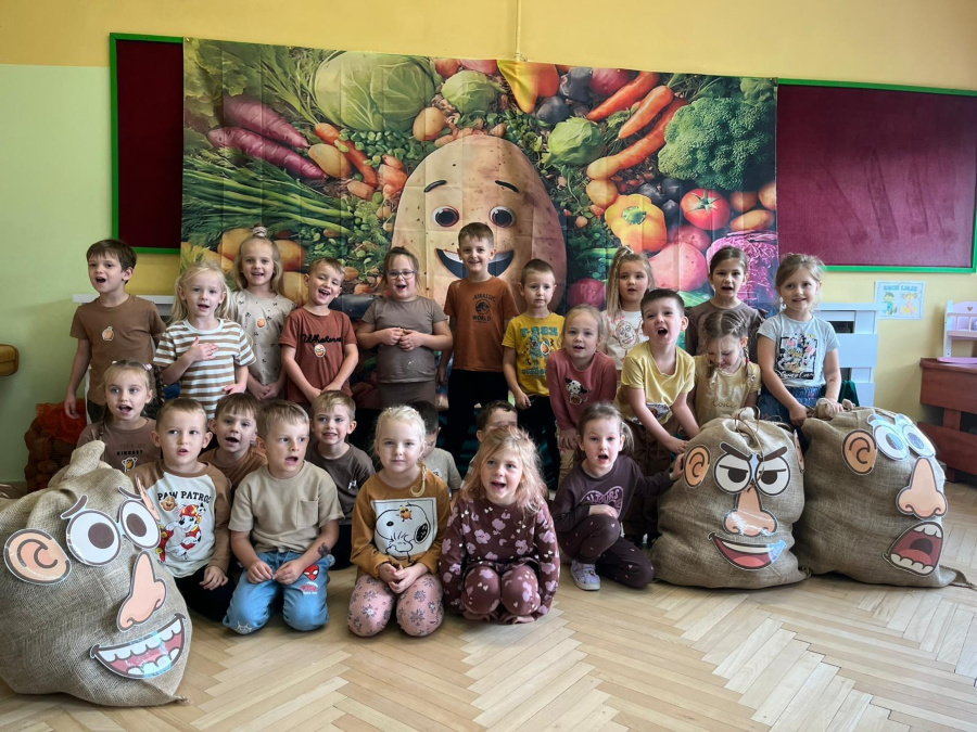 Grupa dzieci pozuje do zdjęcia na tle dekoracji z wizerunkiem dużego ziemniaka otoczonego warzywami. Dzieci ubrane są w brązowe beżowe i żółte stroje. Przed nimi stoją worki ozdobione dużymi