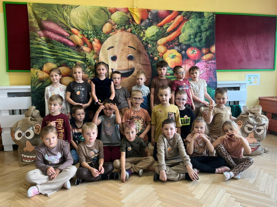 Grupa dzieci pozuje do zdjęcia przed dekoracją przedstawiającą ziemniaka otoczonego warzywami. Dzieci ubrane są w stroje w odcieniach brązu i beżu. Po bokach znajdują się worki 