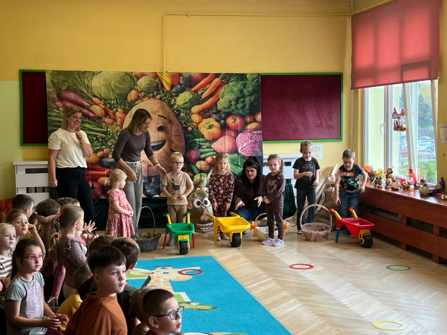 Grupa dzieci stoi w rzędzie z kolorowymi taczkami i koszami. Obok nich znajdują kobiety. Pozostałe dzieci siedzą na dywanie. Tło stanowi kolorowa dekoracja z wizerunkiem ziemniaka