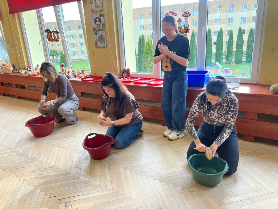 Trzy kobiety klęczą na podłodze trzymając ziemniaki nad miskami obierają ziemniaki. Czwarta kobieta stoi obok klaszcze w ręce. W tle widać duże okna