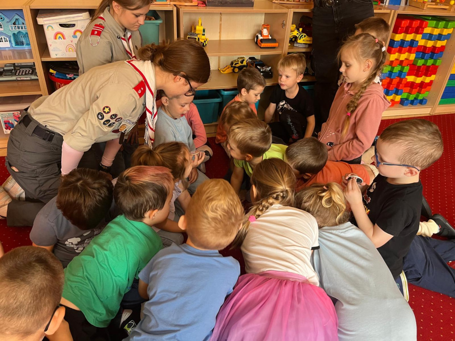 Dzieci siedzą blisko siebie na dywanie skupione wokół dorosłych którzy pokazują im coś ciekawego. W tle widać półki z zabawkami i kolorowymi klockami