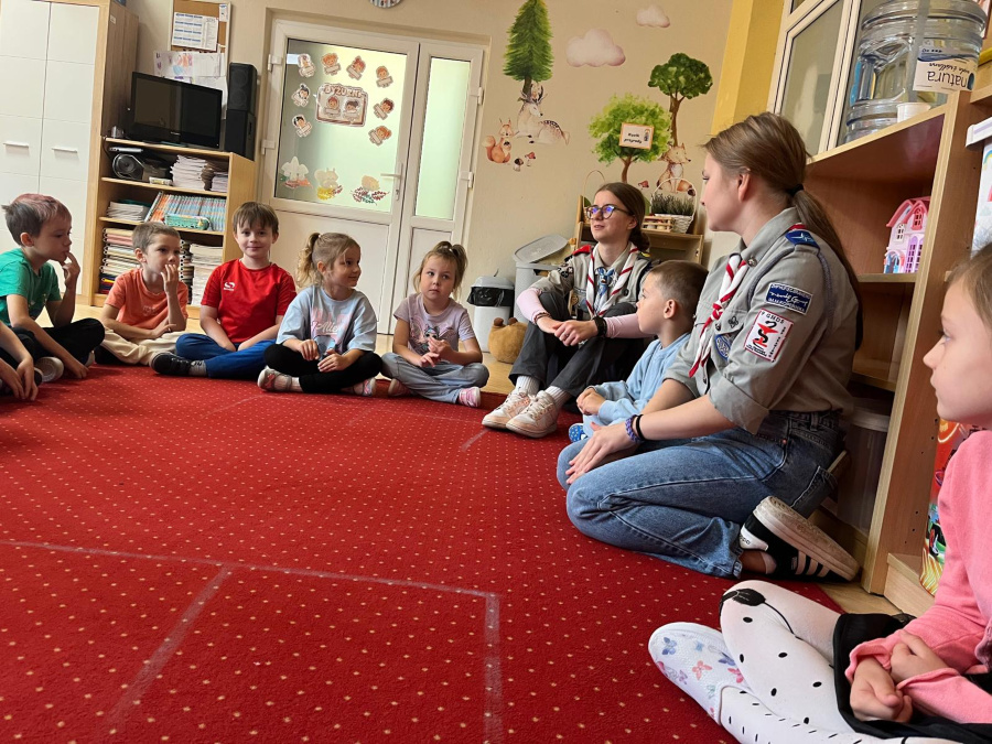 Dzieci siedzą w kręgu na czerwonym dywanie, uważnie słuchając dwóch harcerek w mundurach. W sali widać dekoracje z motywami leśnymi oraz półki z zabawkami i książkami