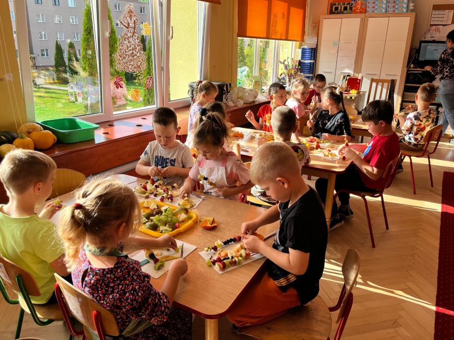 Dzieci przygotowują szaszłyki owocowe przy stolikach w sali przedszkolnej. Każde dziecko ma przed sobą tackę z kawałkami owoców które nadziewa na patyczki