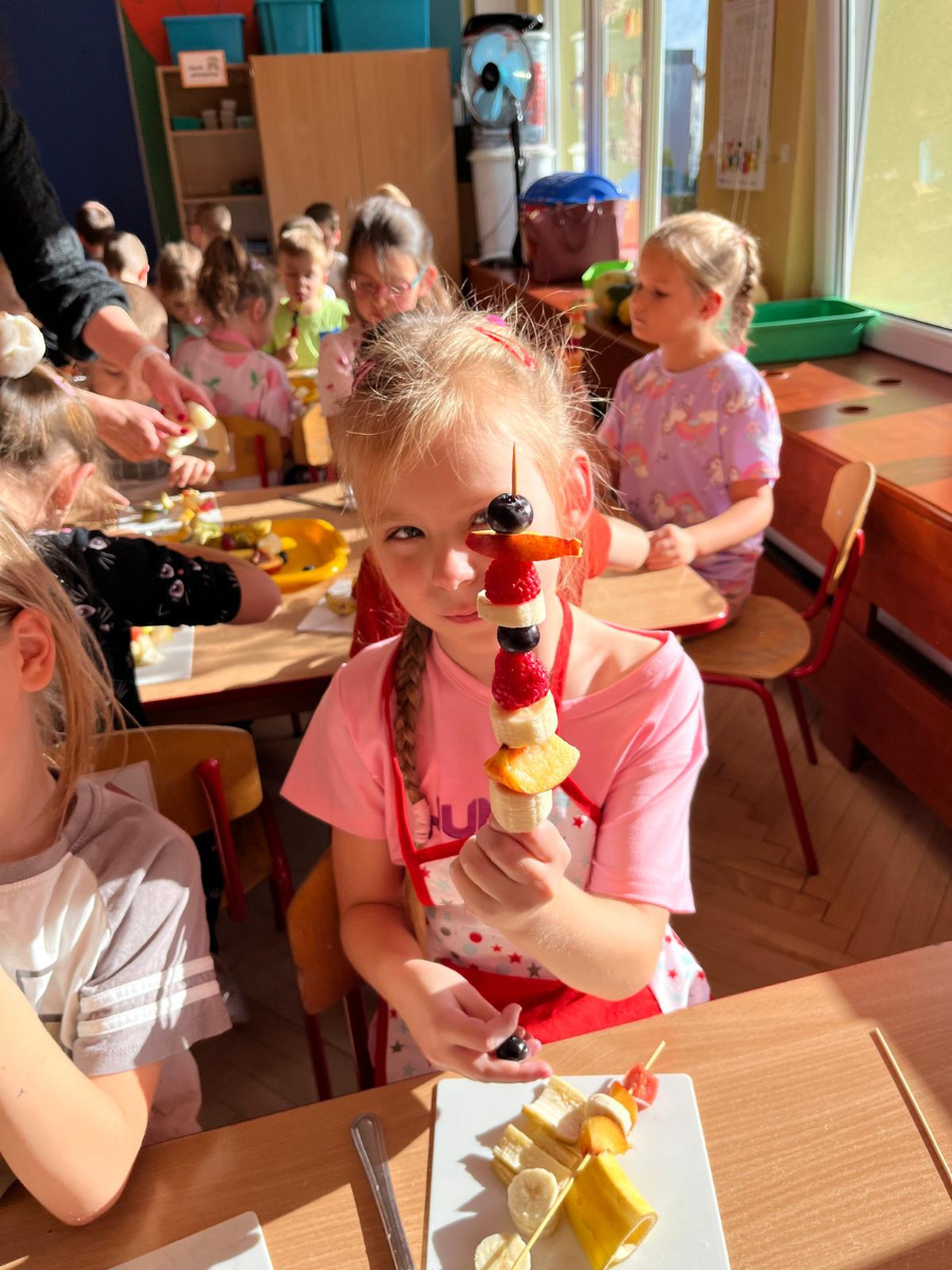 Dziewczynka trzyma przed sobą owocowego szaszłyka. Na patyczku znajdują się kawałki banana truskawki winogrona i brzoskwini. W tle widać inne dzieci zajęte tworzeniem szaszłyków