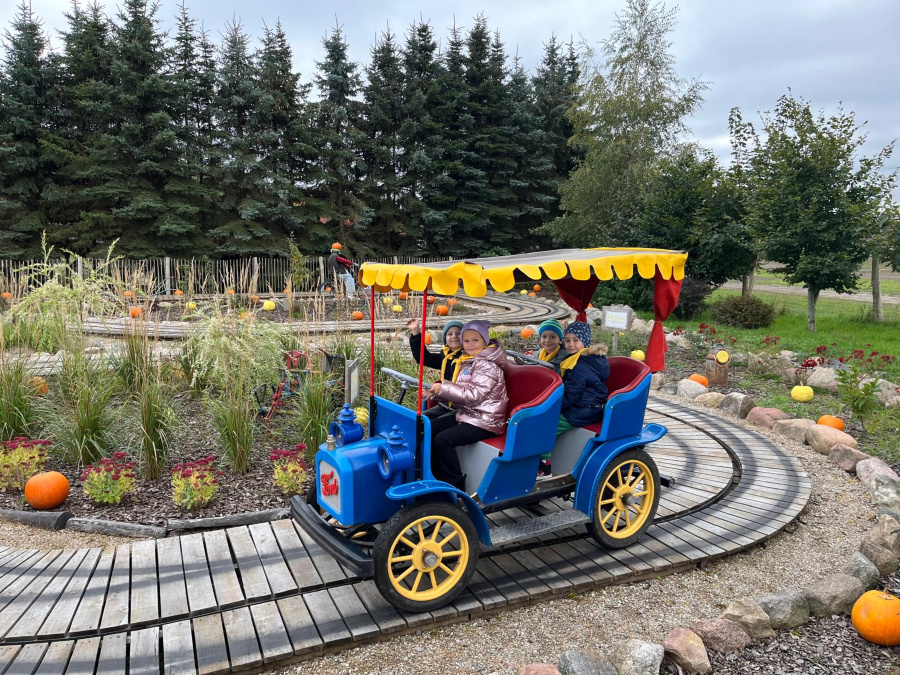 Dzieci jadą niebieskim autkiem w stylu retro po torze kolejki w parku rozrywki. Samochodzik ma żółty dach z czerwonymi zasłonkami a wokół toru rosną trawy i stoją dynie. Dzieci uśmiechają się