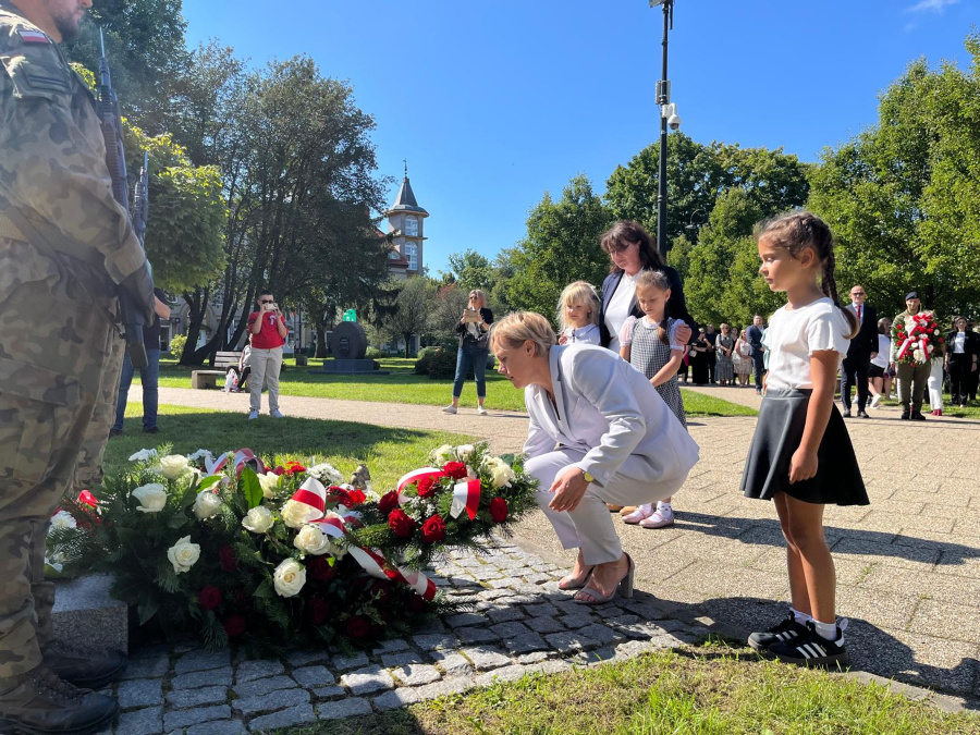 Kobieta w białym garniturze pochyla się aby złożyć wieniec z biało czerwonymi wstęgami przed pomnikiem. Obok niej stoją dzieci i kobieta ubrana na czarno z biał