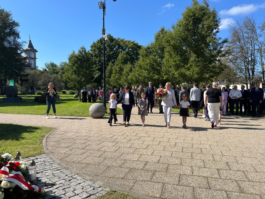 Kobieta w jasnym garniturze niesie wieniec z biało-czerwonymi kwiatami, idąc za rękę z dwiema dziewczynkami. Obok niej podąża kobieta w ciemnym stroju z kolejnym