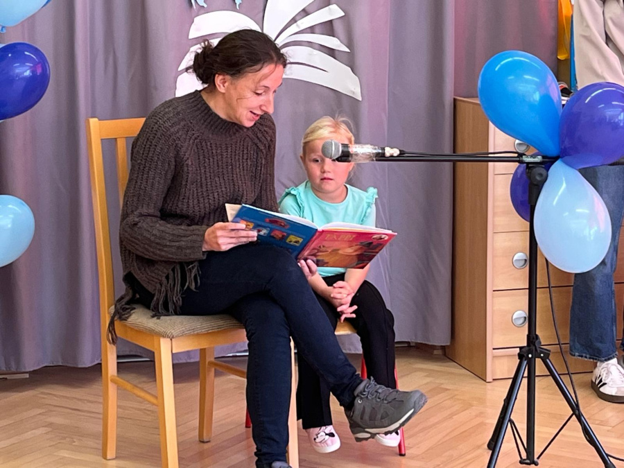 Kobieta siedzi na krześle i czyta książkę a obok niej na małym krzesełku siedzi dziewczynka w jasnoniebieskiej bluzce. Przed nimi ustawiony jest mikrofon ozdobiony balonami