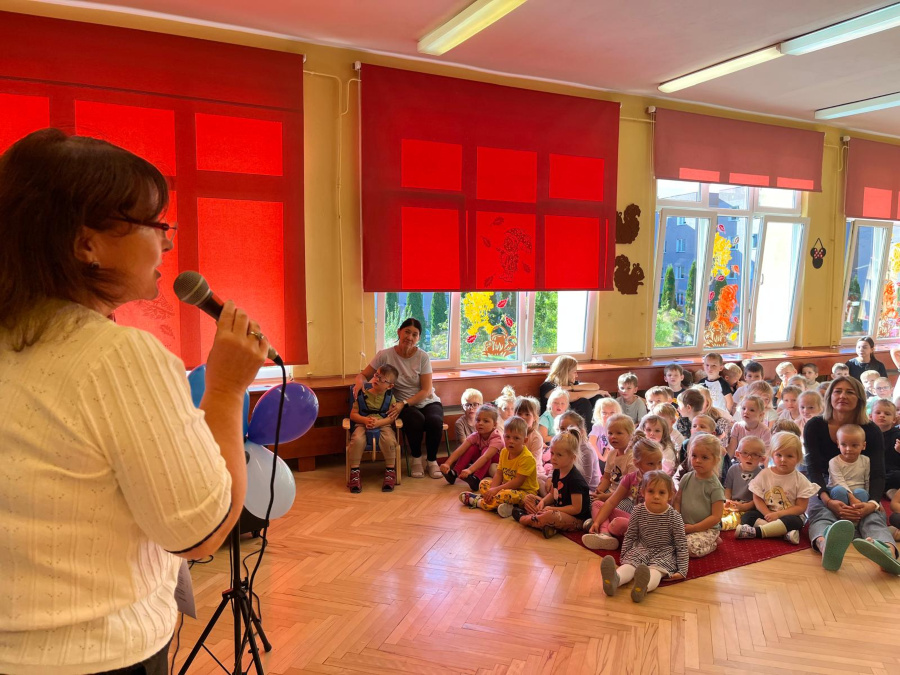 Kobieta trzymająca mikrofon zwraca się do grupy dzieci siedzących na dywanie. W tle widać duże okna ozdobione jesiennymi dekoracjami