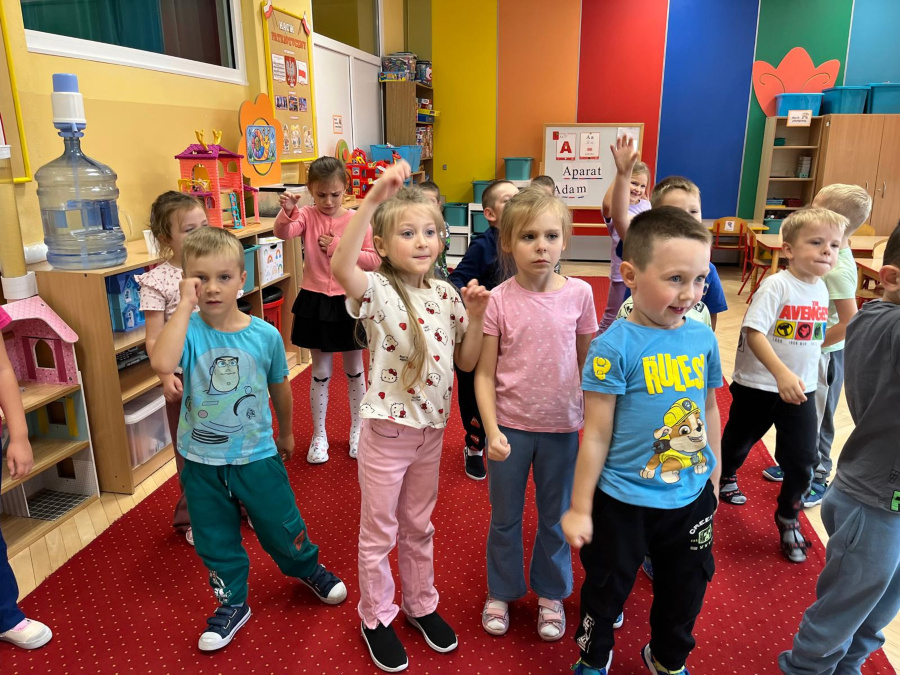 Grupa dzieci stoi na czerwonym dywanie. Są uśmiechnięte niektóre podnoszą ręce do góry inne pokazują gesty dłonią. W tle widać kolorowe ściany zabawki na półkach oraz stoliki