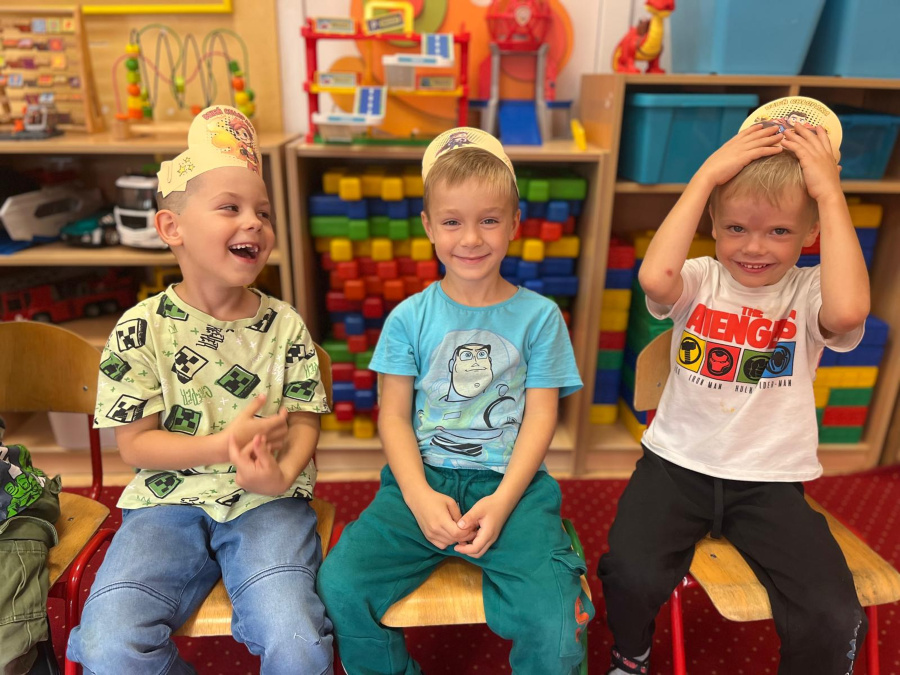 Trzech chłopców siedzi obok siebie na krzesełkach. Każdy ma na głowie papierową opaskę a na twarzach widać radość i uśmiechy. W tle stoją kolorowe klocki oraz zabawki poukładane na półkach