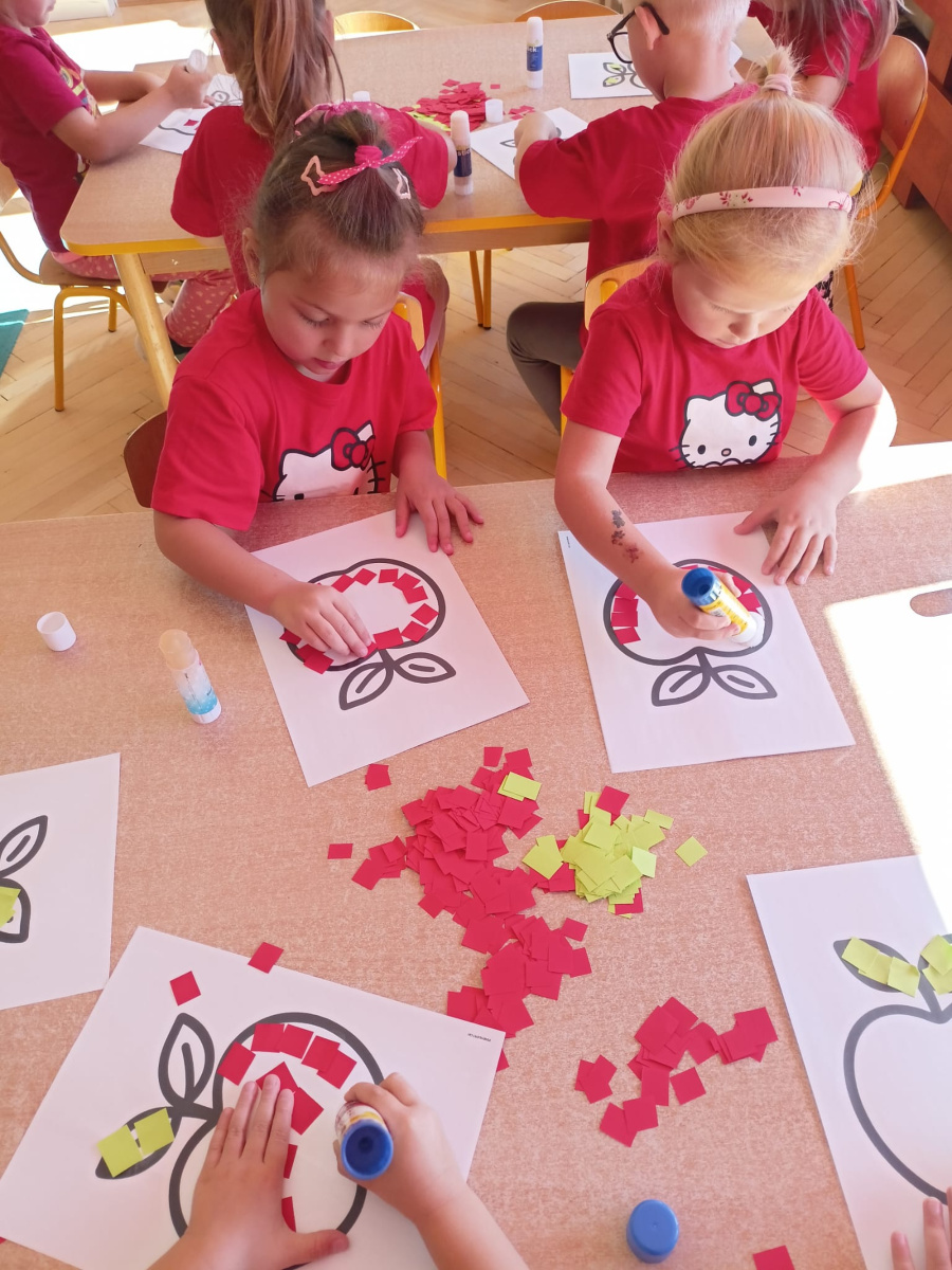 Dzieci siedzą przy stoliku. Każde dziecko ma przed sobą kartkę z konturem jabłka które wykleja kolorowymi kwadracikami z czerwonego i zielonego papieru. Na środku stołu leżą kolorowe papierki