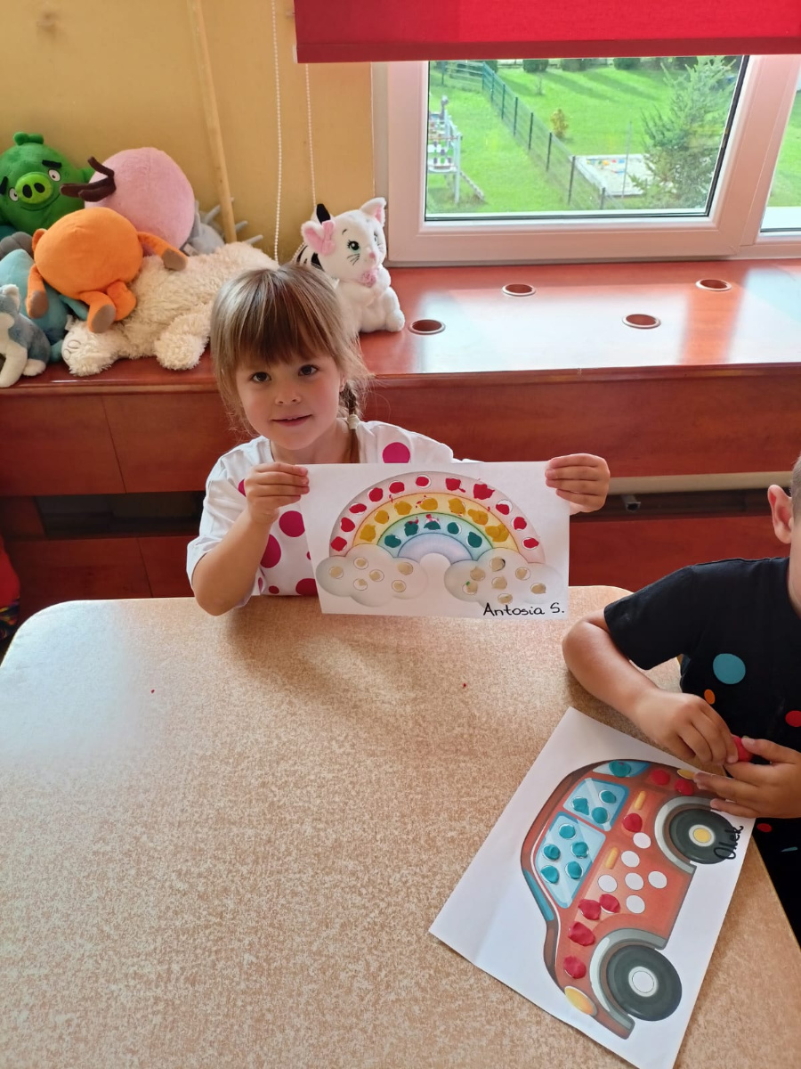 Dziewczynka siedzi przy stoliku i trzyma przed sobą kartkę tęczę z chmurkami ozdobioną kolorowymi naklejkami. Na stole leży także obrazek samochodu który został udekorowany w podobny sposób