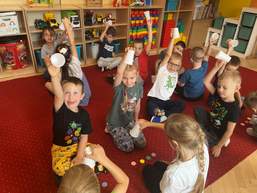 Grupa dzieci siedzi na czerwonym dywanie. Każde z nich trzyma w ręku biały plastikowy kubeczek, większość podnosi je wysoko do góry z uśmiechem. W tle widać kolorowe półki z zabawkami i klockami