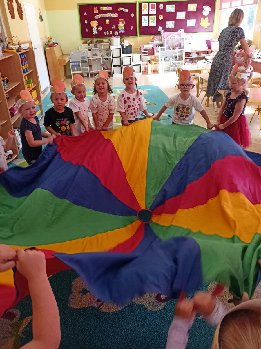 Grupa dzieci stoi w kole i trzyma dużą kolorową chustę w pasy czerwone żółte zielone i niebieskie. Na głowach mają papierowe opaski z pomarańczowymi elementami a na ich twarzach widać uśmiechy