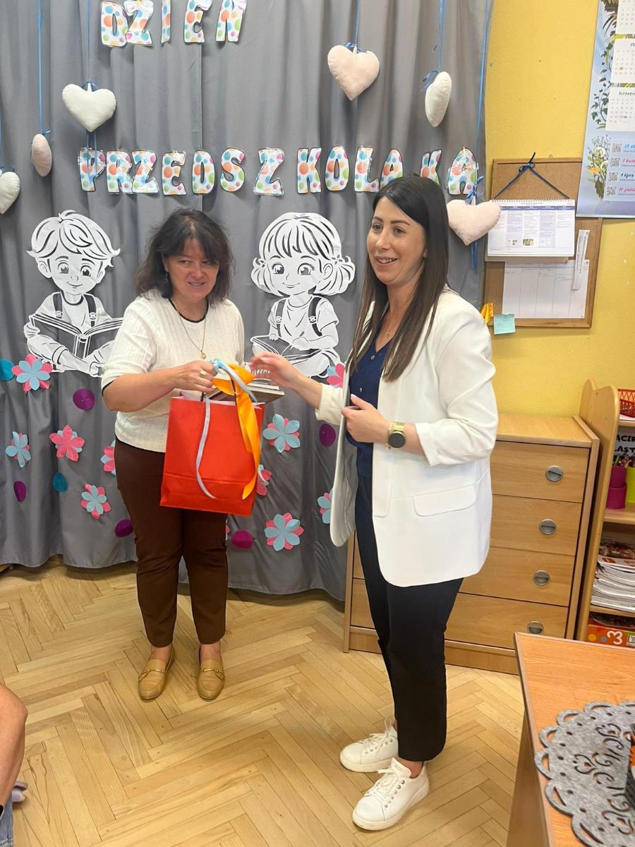 Dwie kobiety stoją przed dekoracją z napisem Dzień Przedszkolaka. Jedna z nich trzyma czerwony prezentowy worek ozdobiony kolorową wstążką a druga właśnie go wręcza. Obie uśmiechają się