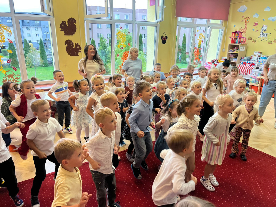 Grupa dzieci bierze udział w zabawie ruchowej. Dzieci stoją na czerwonym dywanie, uśmiechają się i energicznie wykonują ruchy. W tle widać duże okna ozdobione kolorowymi jesiennymi dekoracjami