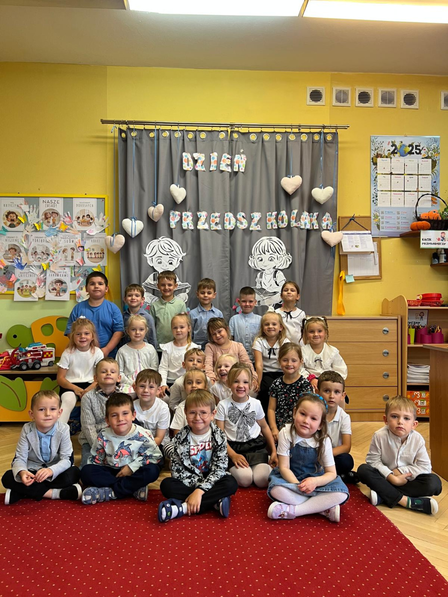 Grupa dzieci pozuje do wspólnego zdjęcia. Siedzą i stoją na tle dekoracji z napisem Dzień Przedszkolaka ozdobionej serduszkami i rysunkami dzieci