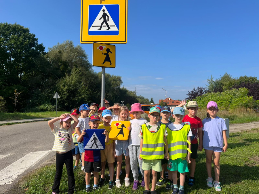 Grupa dzieci stoi przy drodze obok znaków drogowych informujących o przejściu dla pieszych. Dwoje dzieci trzyma w rękach plansze przedstawiające symbole przejścia dla pieszych