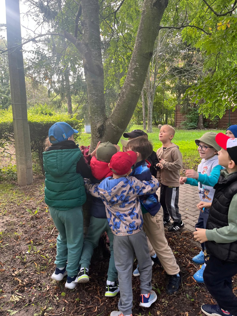 Grupa dzieci na świeżym powietrzu obejmuje pień drzewa. Dzieci stoją blisko siebie przytulając się do drzewa lub trzymając je ramionami. Ubrane są w kolorowe bluzy kamizelki i czapki