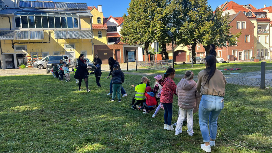 Dzieci biorą udział w przeciąganie liny na trawie. Uczestnicy stoją w dwóch drużynach. Obok stoją kobiety które dopingują i czuwają nad przebiegiem zabawy