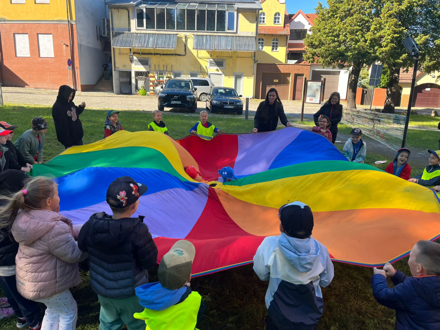 Dzieci biorą udział w zabawie z kolorową chustą animacyjną na świeżym powietrzu. Trzymają jej brzegi,  na materiale leżą piłeczki i maskotka