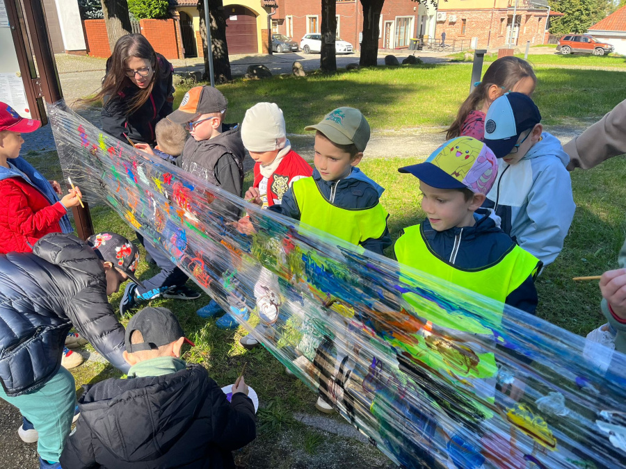 Dzieci malują farbami na przezroczystej folii rozwieszonej między słupkami. Tworzą barwne kompozycje wykorzystując różne kolory i kształty. Obok czuwa nauczycielka
