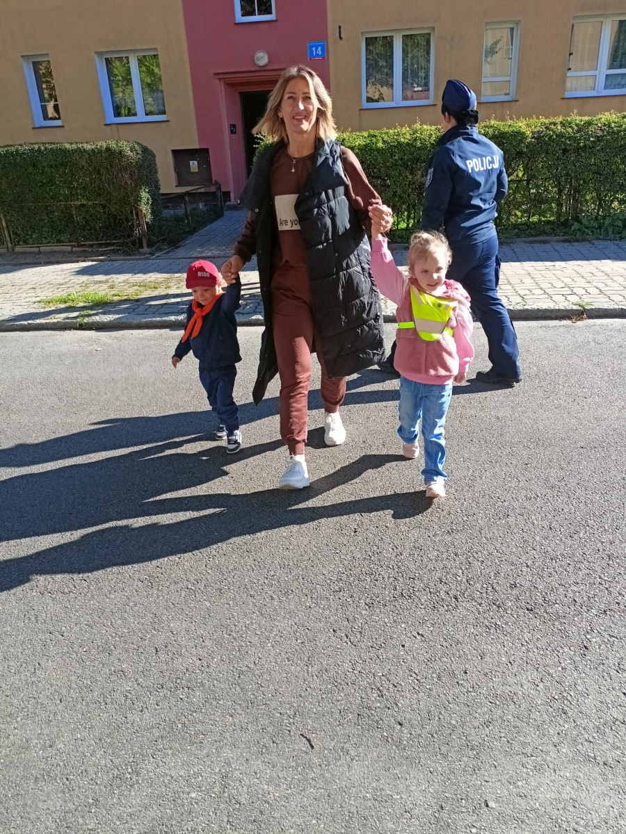 Kobieta prowadzi dwójkę dzieci przez ulicę trzyma ich za ręce. Dziewczynka ma na sobie odblaskową kamizelkę a chłopiec czerwony daszek i pomarańczową chustę. W tle widać policjantkę oraz budynek