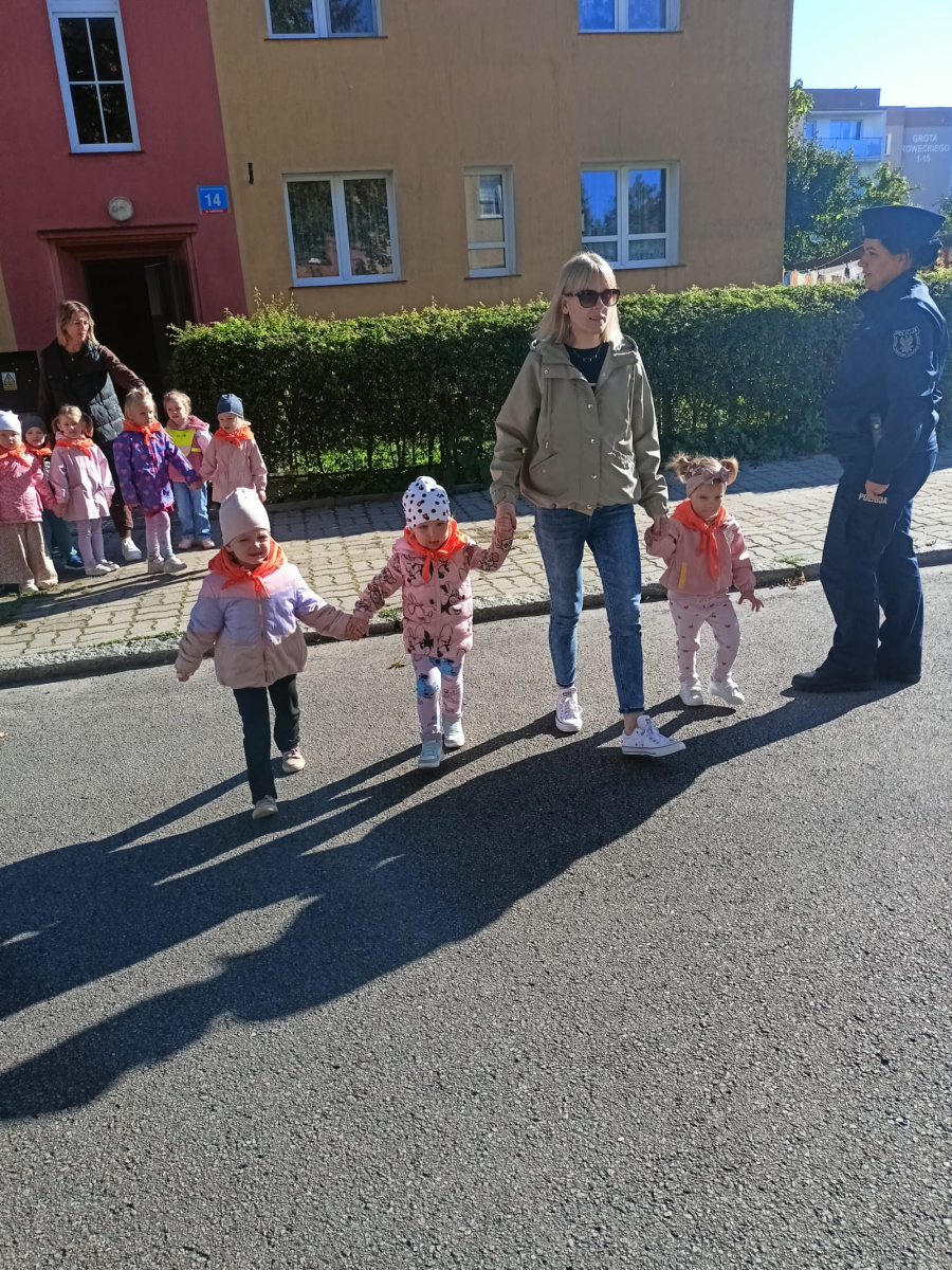 Kobieta w okularach przeciwsłonecznych prowadzi za ręce troje dzieci przez jezdnię. Wszystkie maluchy mają pomarańczowe chusty pod szyją a w tle stoi policjantka