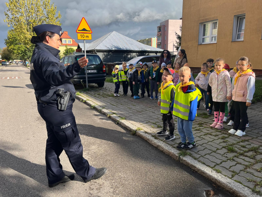 Policjantka w mundurze stoi na ulicy i gestykuluje. Przed nią, na chodniku, ustawiona jest grupa dzieci w odblaskowych kamizelkach i z żółtymi chustami na szyjach. Obok stoją kobiety