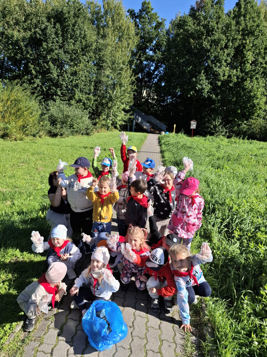 Grupa dzieci stoi i kuca na chodniku w otoczeniu zieleni. Mają na dłoniach jednorazowe rękawiczki a przed nimi leży niebieski worek ze zebranymi śmieciami. Część dzieci ma na szyjach czerwon