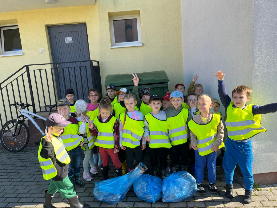 Grupa dzieci w odblaskowych, żółtych kamizelkach stoi razem na tle budynku. Dzieci uśmiechają się i pokazują gesty dłoni, niektóre unoszą ręce w górę. Przed nimi leżą trzy niebieskie worki