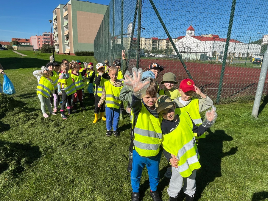 Grupa dzieci w żółtych kamizelkach odblaskowych stoi na trawie przy ogrodzeniu boiska. Kilkoro dzieci w rękawiczkach jednorazowych pokazuje dłonie w stronę aparatu